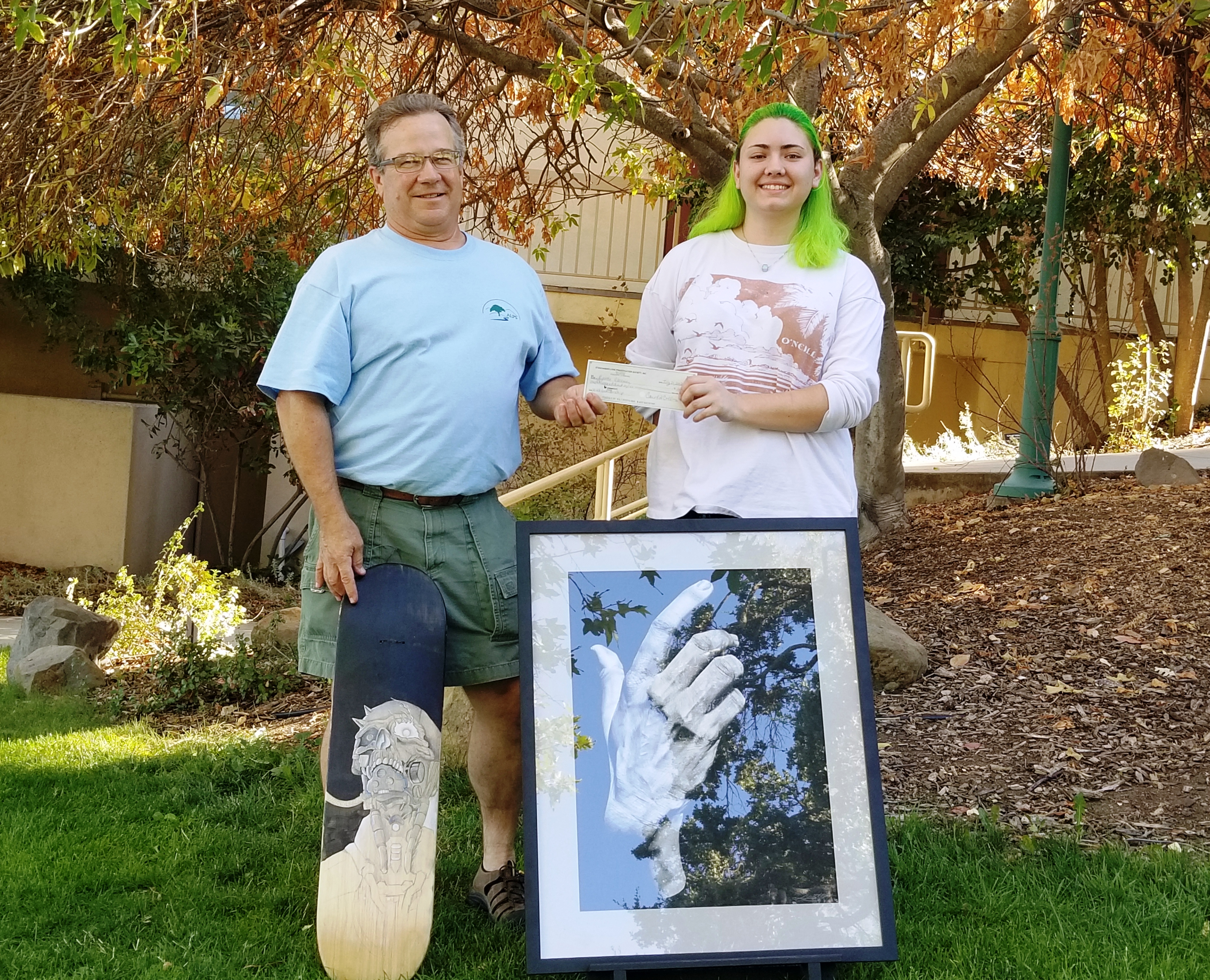 Dani Bloomer receiving scholarship check from Mike Orvis at the July board meeting  (in the foreground are two examples of Dani's art)  